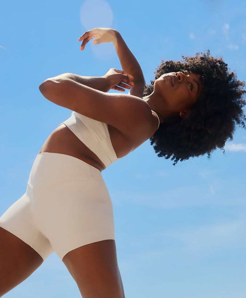 Woman in a sports bra and shorts against a blue sky with sunlight. Female athlete stretches outdoors with arms raised in the sun.