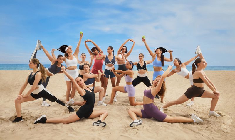 Group of diverse women in activewear stretches and poses with tennis equipment on a sandy beach with a blue sky backdrop.