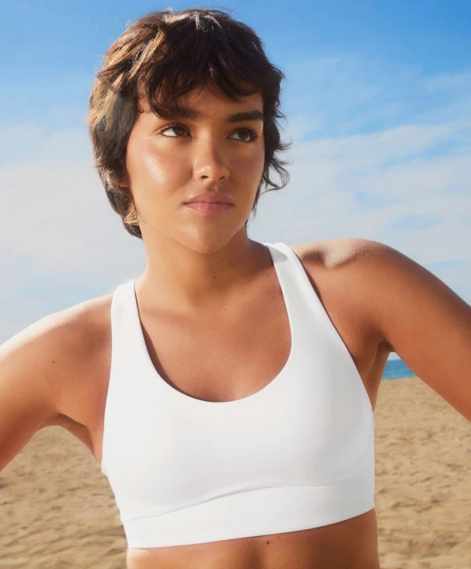 Woman in a white sports bra stands on a beach on a sunny day with a blue sky and ocean in the background.