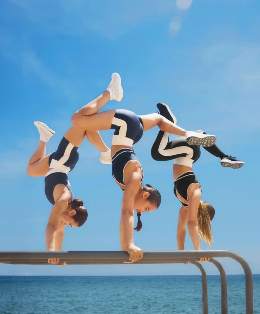 Three adult females in sports attire perform handstand yoga poses on a metal bar outdoors against a blue sky and ocean backdrop on a sunny day.