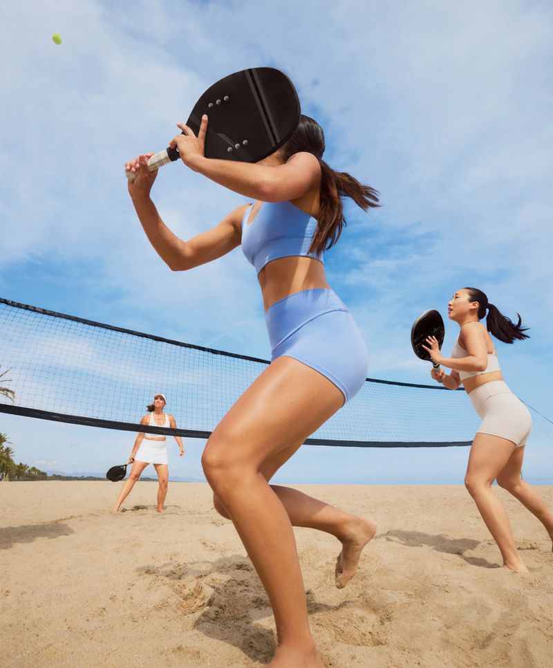 Three women playing beach tennis with racquets on a sunny day.  A black net is visible, with trees and blue sky in the background.
