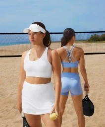 Two women posing with paddle rackets on a sandy beach court wearing white and light blue sportswear sets by a net.