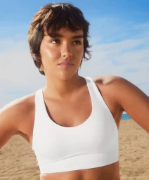 Woman with short dark hair wearing a white sports bra standing on a sunny sandy beach with blue sky.