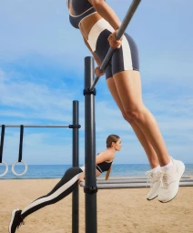 Two women working out on outdoor calisthenics bars in sportswear on a sandy beach with the ocean behind them.