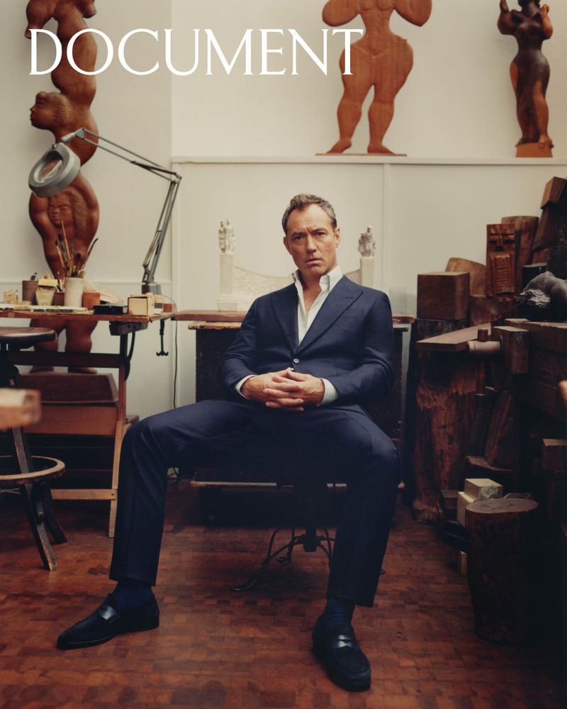 Jude Law seated in an art studio, wearing a blue suit, with wooden sculptures and tools in the background, and the word DOCUMENT on top.