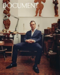 Jude Law seated in a dark navy tailored suit and open-collar white shirt inside an artist studio surrounded by wooden sculptures.