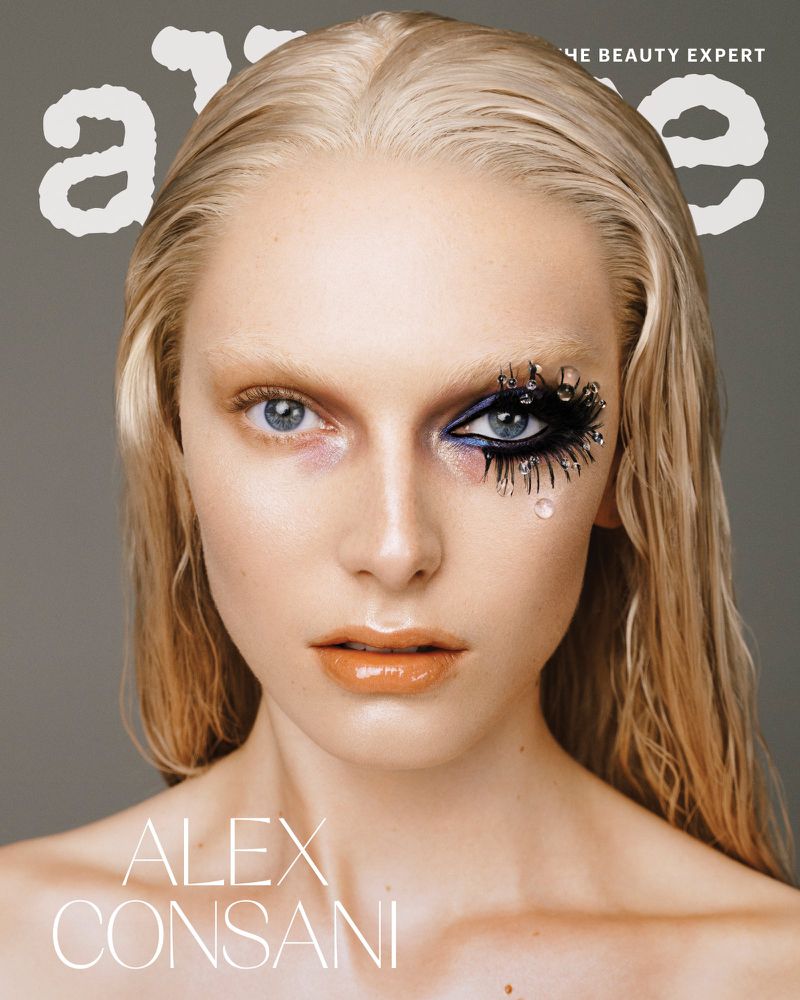 Close-up beauty shot of a blond woman with creative black eye makeup, glossy lips, and grey background for a magazine cover.