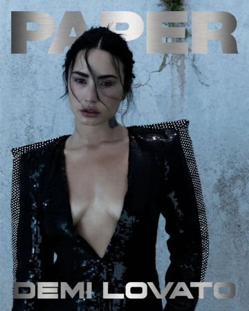 Demi Lovato in a plunging black sequin blazer with sharp structured shoulders against a weathered concrete backdrop.