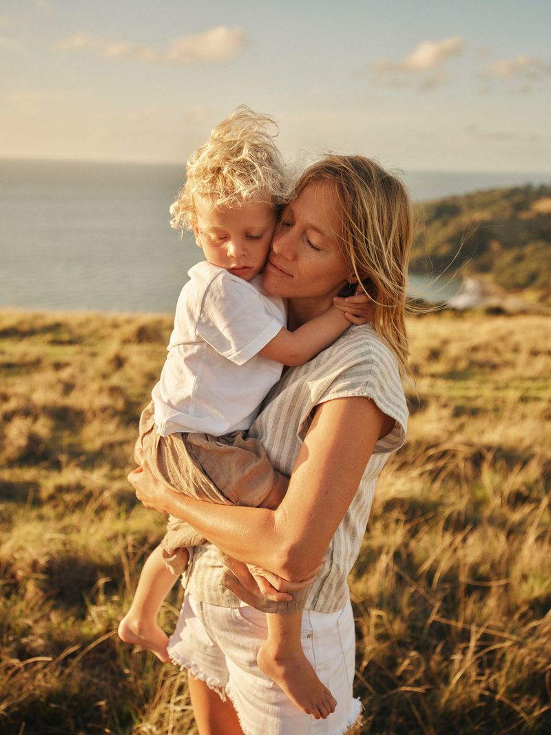A mother holds her child in a grassy field near the ocean under soft sunlight, creating a warm and intimate outdoor moment.