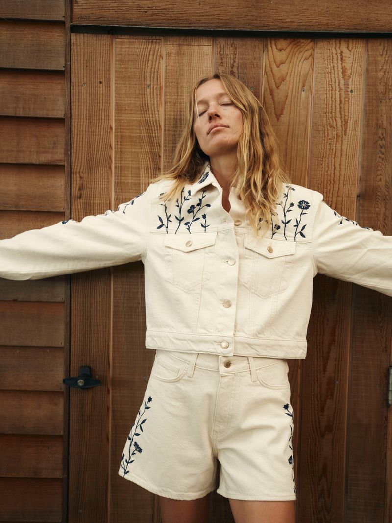 Blonde woman in embroidered denim jacket and shorts poses against a wooden wall, eyes closed, arms outstretched in bright outdoor lighting.