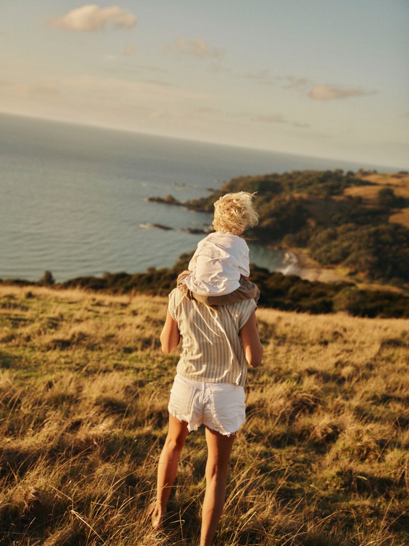A person carries a child on their shoulders walking through a grassy field by the ocean at sunset, creating a serene and joyful scene.