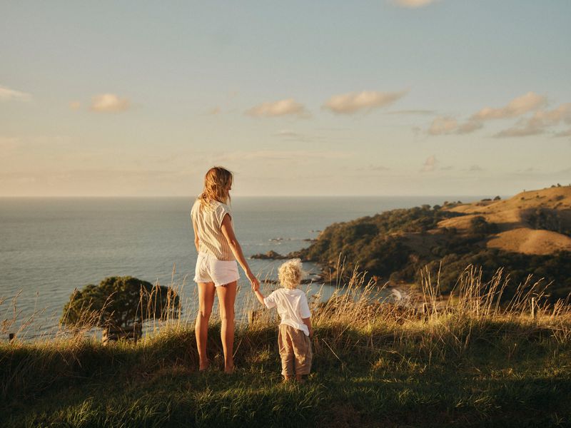 Woman and child holding hands on a grassy coastal hill overlooking the ocean at sunset