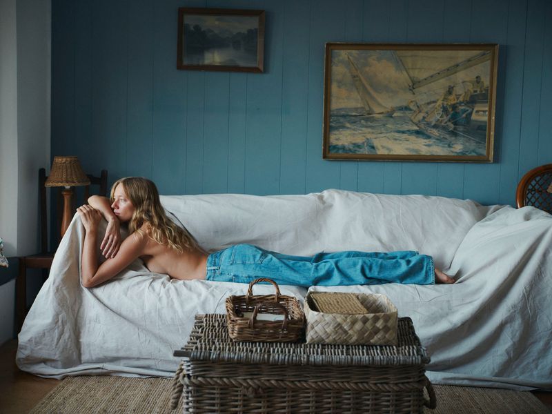 Blonde woman lies on white sofa in blue jeans, indoors. Paintings hang on blue walls with wicker table and baskets nearby.
