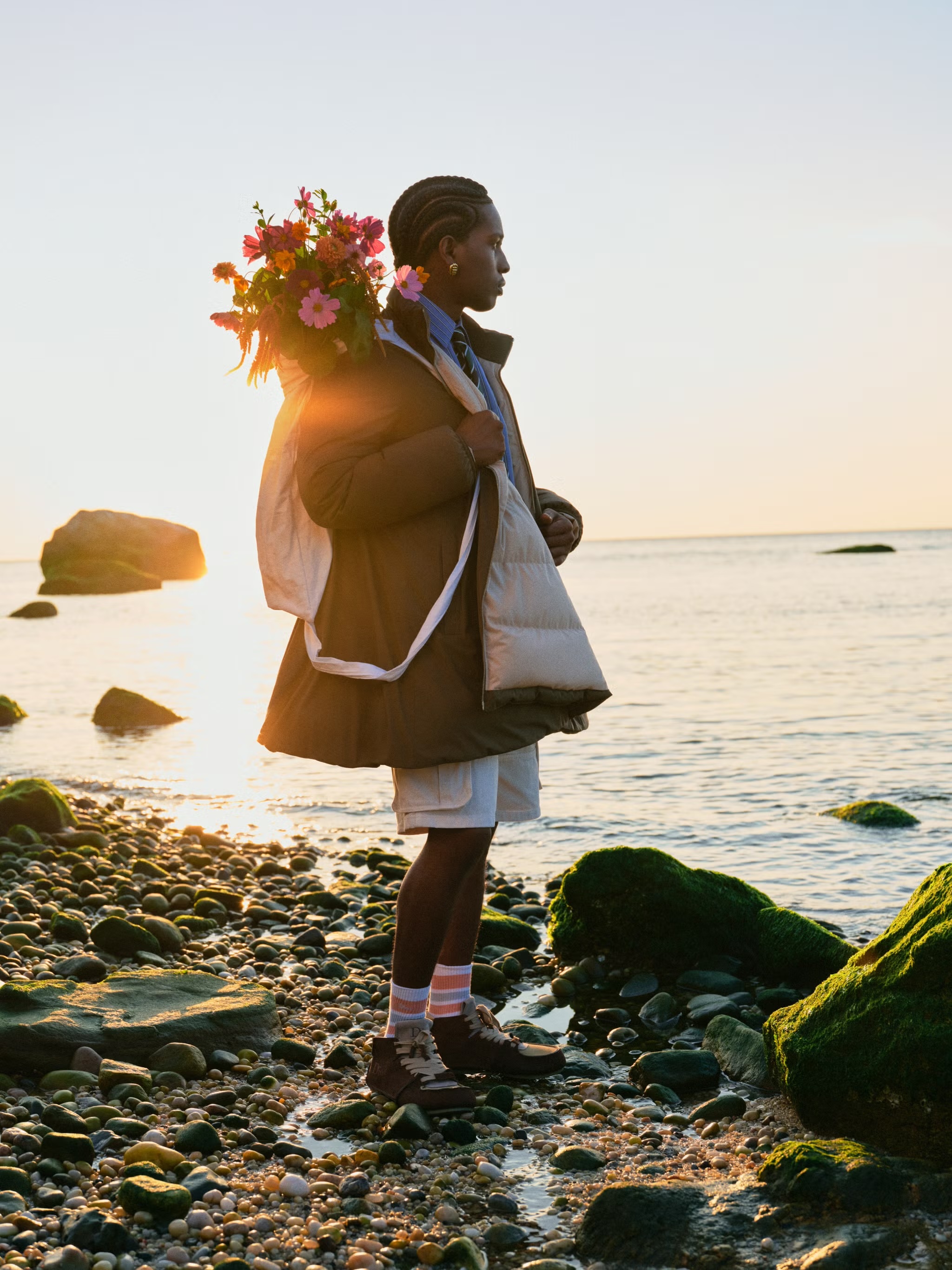 Black man holding a large bouquet of colorful flowers on a rocky shore at sunset