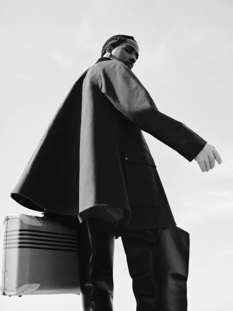 Monochrome fashion portrait of a Black man with braids wearing a cape coat and holding a suitcase against a bright, overcast sky.
