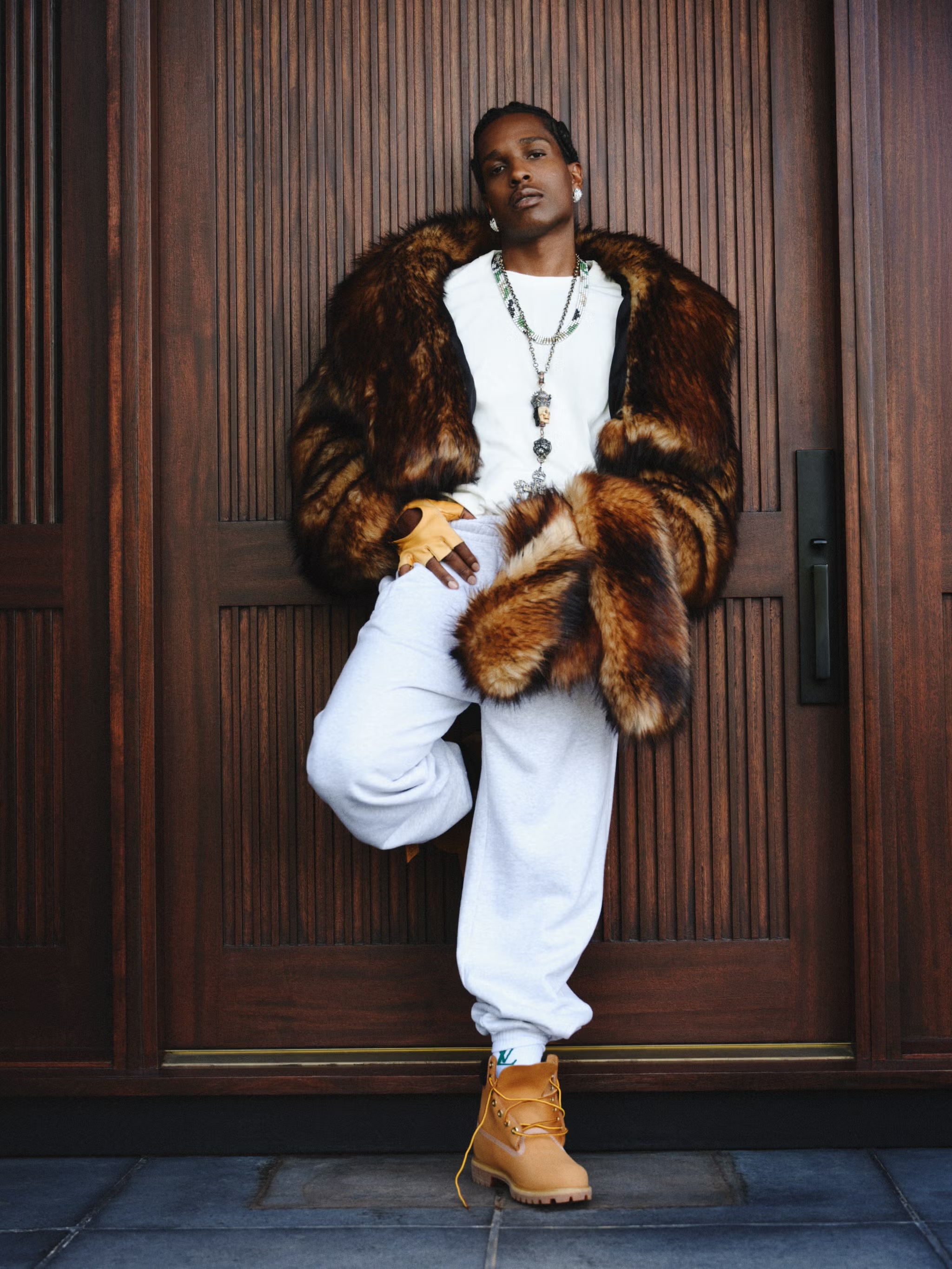 A man in a fur coat leans against a dark wood door wearing a white t-shirt, sweatpants, boots and multiple necklaces. He has a serious expression.