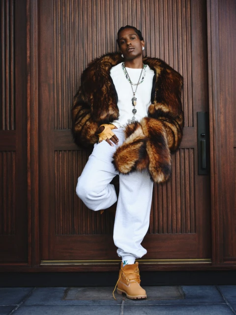 A man in a fur coat leans against a dark wood door wearing a white t-shirt, sweatpants, boots and multiple necklaces. He has a serious expression.