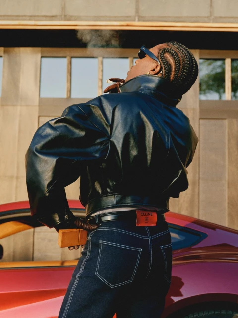 Stylish adult with braids, sunglasses and leather jacket exhales smoke near a red sports car with a concrete building backdrop.