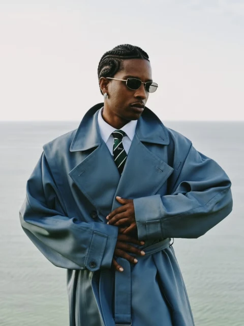 A stylish African American man with cornrows, sunglasses, and a trench coat poses against a light sky with a hint of a body of water.