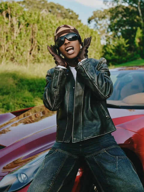 A male model leans against a car, wearing a black leather jacket, dark denim jeans, and sunglasses against a nature background.
