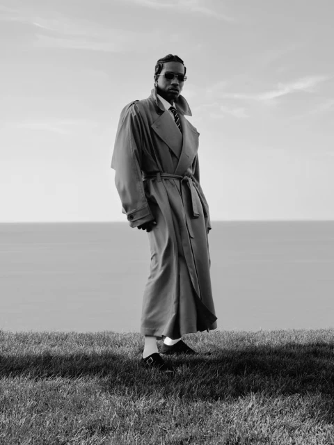 Black and white portrait of an African American man in a trench coat standing on a grassy hill overlooking the ocean, under a cloudy sky.