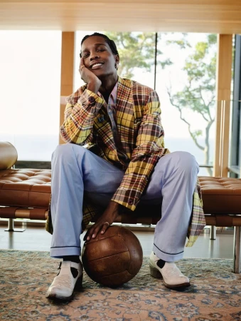 Adult male sitting on a leather bench, wearing a yellow plaid jacket and light blue pants while holding a brown soccer ball.