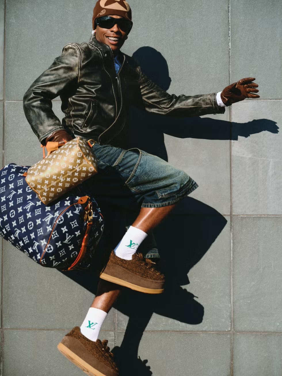 Smiling adult male in streetwear attire jumps in front of a grey tiled wall with designer luggage and shoes.