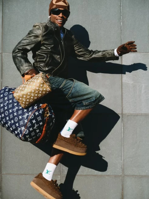 Smiling adult male in streetwear attire jumps in front of a grey tiled wall with designer luggage and shoes.