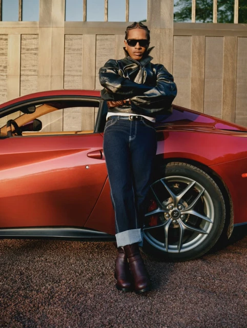 A man in a leather jacket poses with a red Ferrari, showcasing automotive style and fashion against a light-toned architectural backdrop.