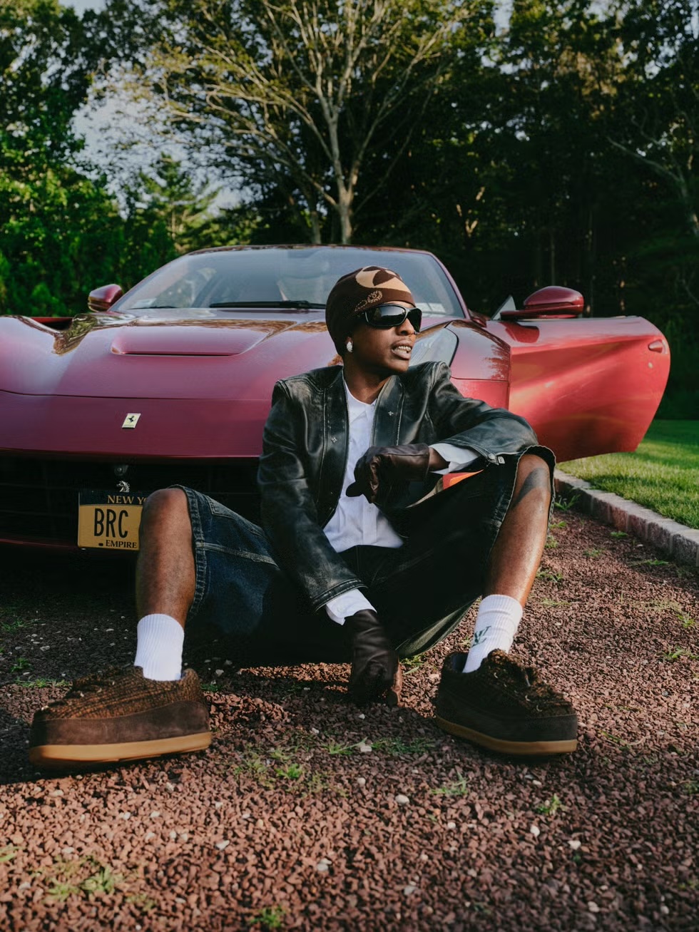 Man sits in front of a red Ferrari on a gravel surface, wearing a dark jacket, beanie, and sunglasses, with trees in the background.