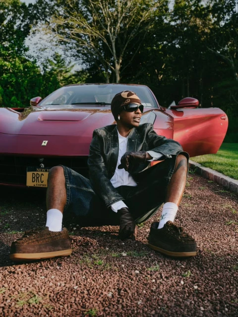 Man sits in front of a red Ferrari on a gravel surface, wearing a dark jacket, beanie, and sunglasses, with trees in the background.