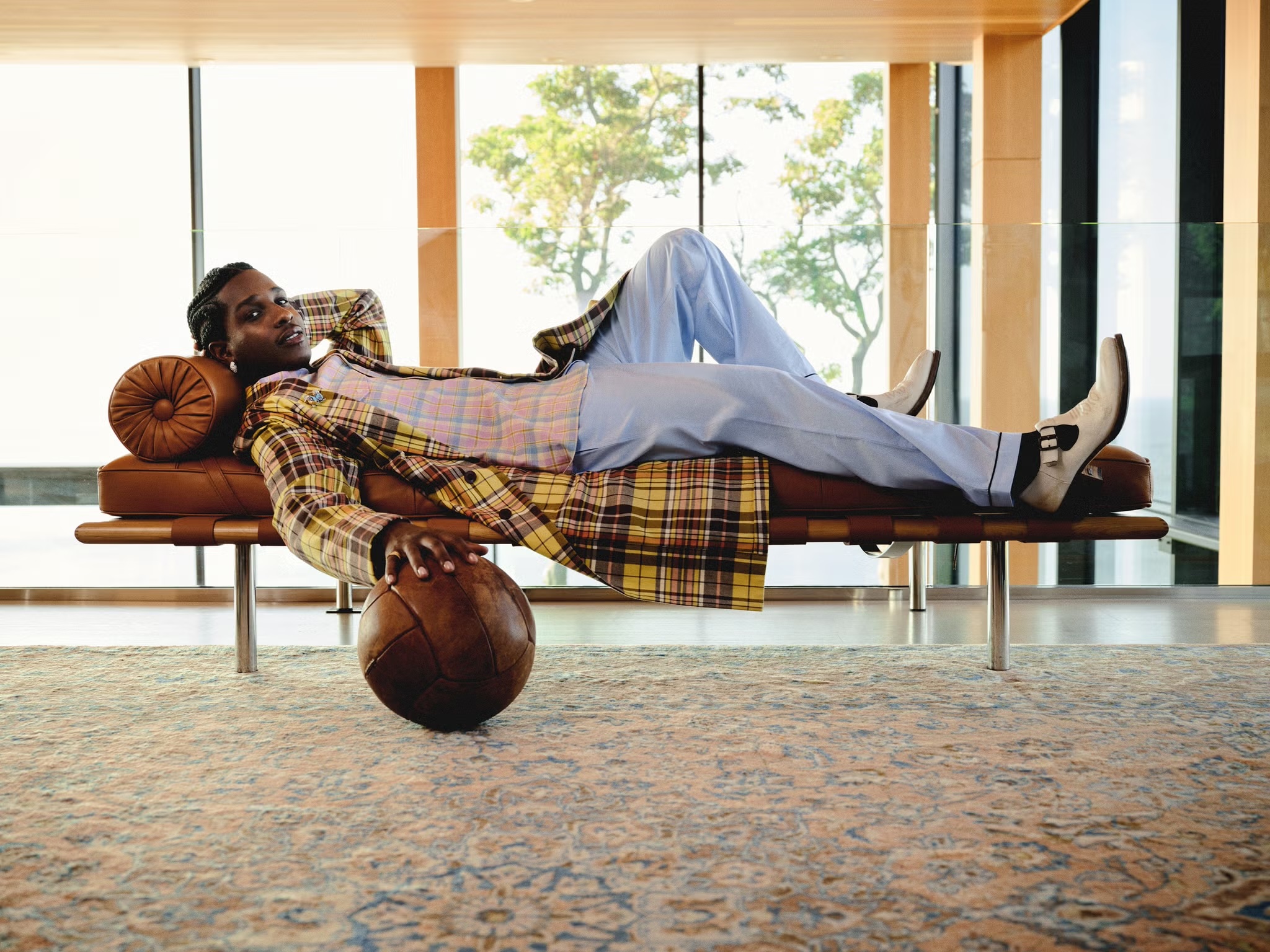 Adult male relaxing on a modern leather daybed in a bright, minimalist room with a vintage soccer ball and large windows overlooking trees.