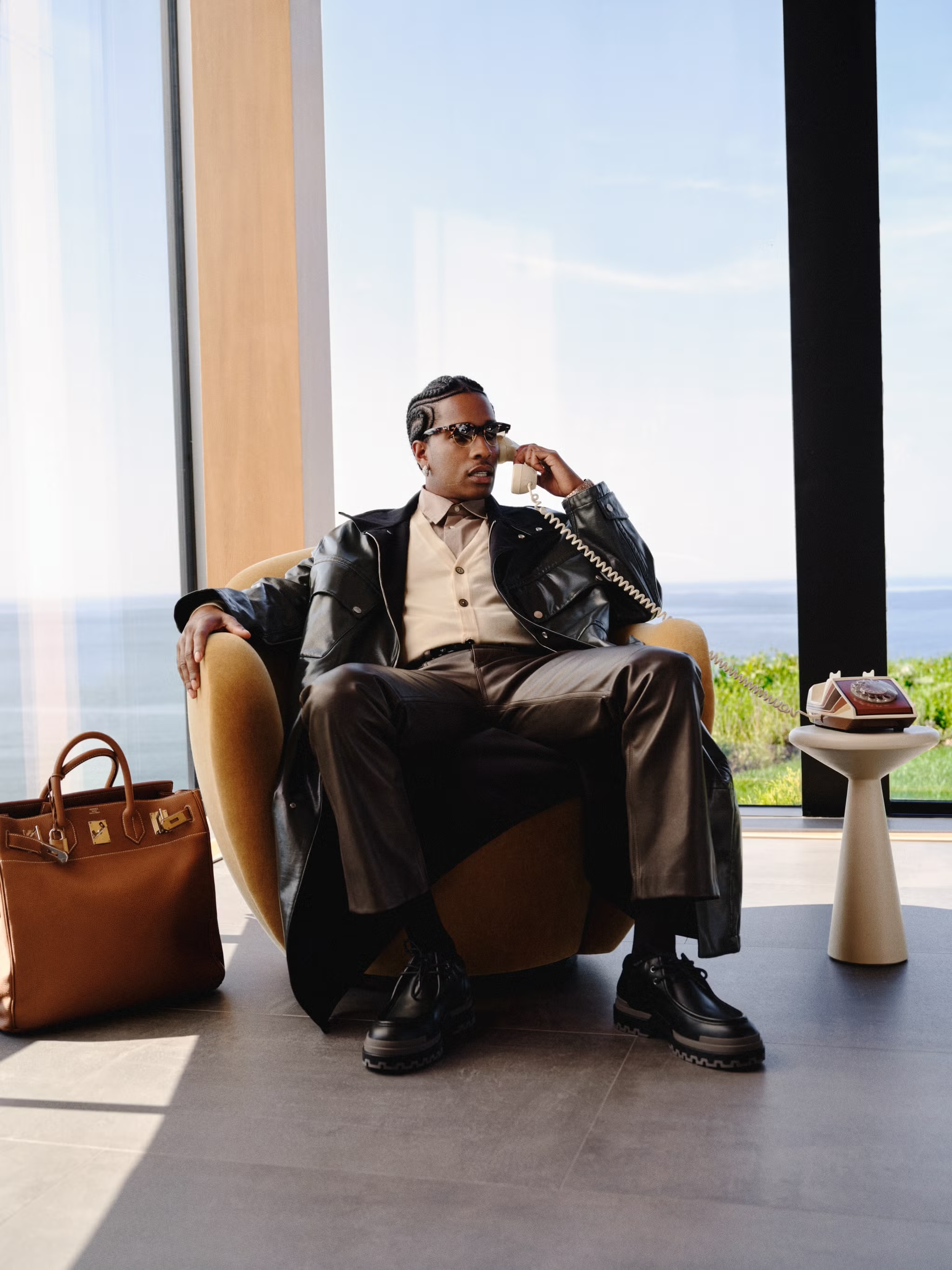 Man in a leather jacket sitting in an armchair, talking on the phone in a modern house with a tan leather bag beside him.