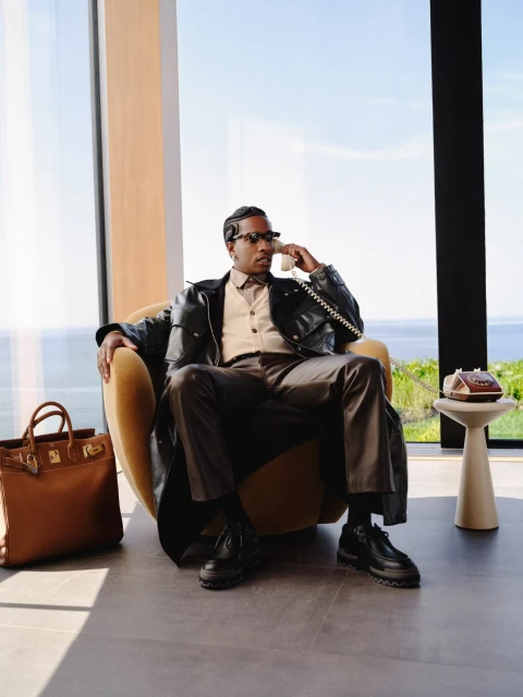 Man in a leather jacket sitting in an armchair, talking on the phone in a modern house with a tan leather bag beside him.