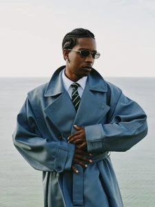 Man wearing a blue trench coat, white shirt, striped tie, and sunglasses against a hazy coastal horizon background.