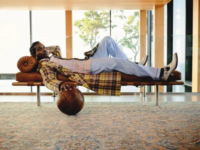 Man reclining on a leather daybed holding a leather soccer ball, wearing a plaid coat and light blue trousers.