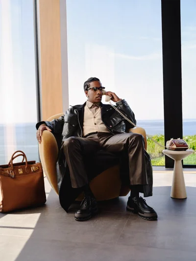 A man wearing a black leather coat and sunglasses seated in a mustard armchair making a phone call, with a brown leather bag beside him