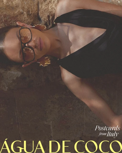 A woman poses with sunglasses and gold earrings on a stone surface while wearing a black top; the photo has a warm, filtered aesthetic.