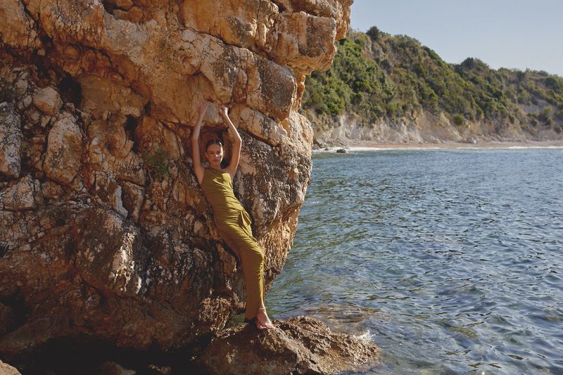 Woman in olive green dress stands barefoot against a tan rock formation with blue ocean and green hillside visible in the background.