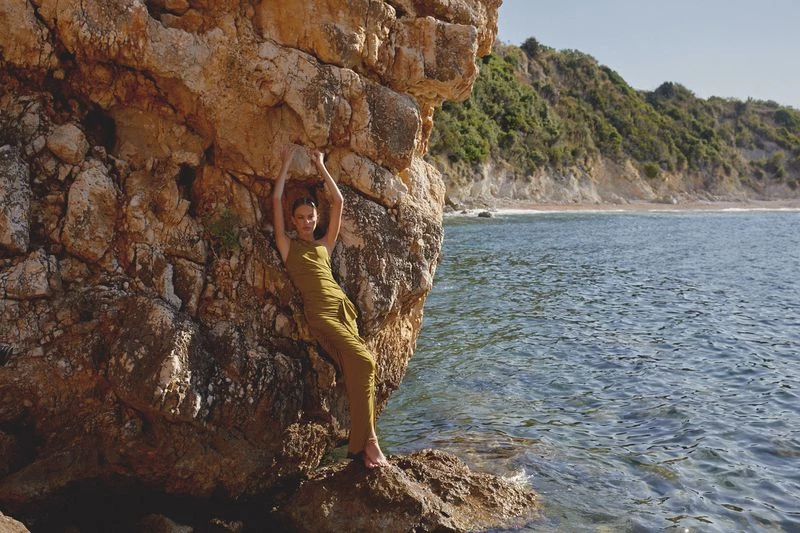 Woman in olive green dress stands barefoot against a tan rock formation with blue ocean and green hillside visible in the background.