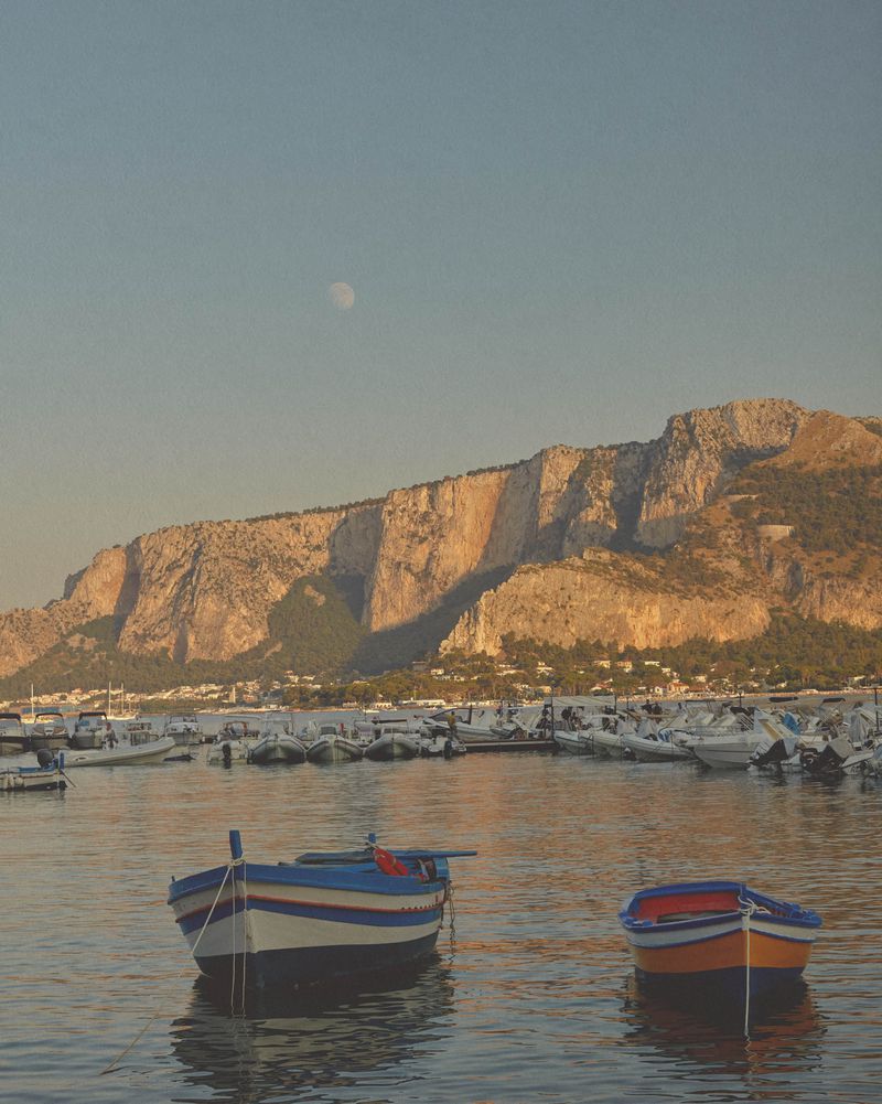 Scenic view of two small boats floating in a calm harbor with a rocky mountain range backdrop under a clear sky, early morning light.