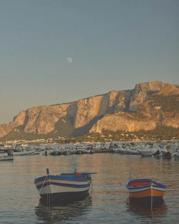 Scenic view of two small boats floating in a calm harbor with a rocky mountain range backdrop under a clear sky, early morning light.