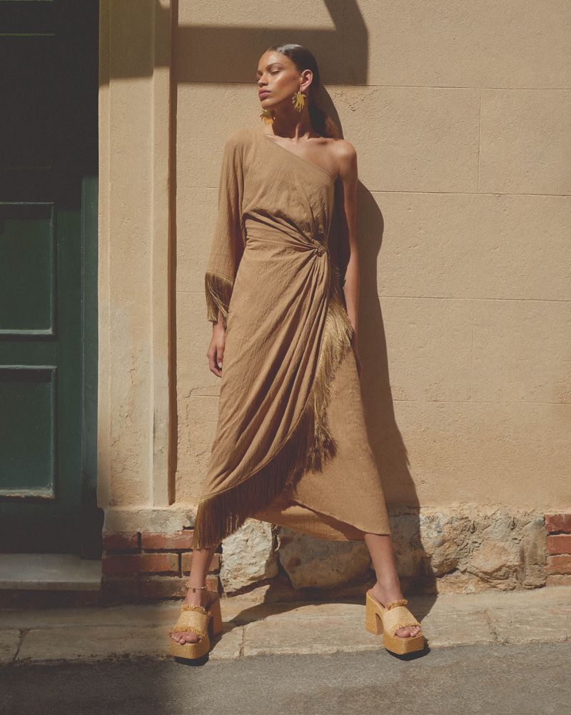 A woman poses in a beige one-shoulder fringe dress and platform sandals in front of a textured wall and green door for a fashion shoot.