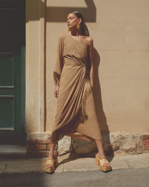 A woman poses in a beige one-shoulder fringe dress and platform sandals in front of a textured wall and green door for a fashion shoot.