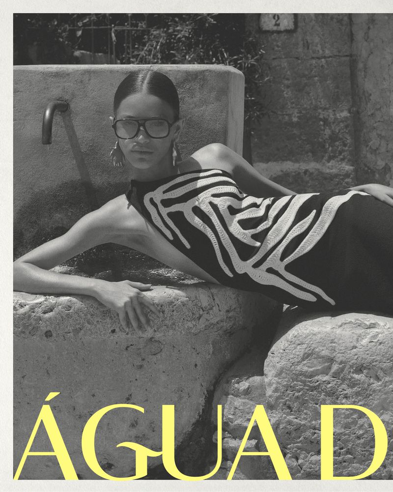 A woman with sunglasses and zebra-striped top reclines next to a stone fountain in a fashion-style black and white photograph with yellow text.