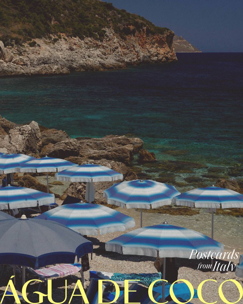 Beach umbrellas line the sandy shore of a rocky Italian beach, with turquoise water and cliffs in the background on a sunny day.