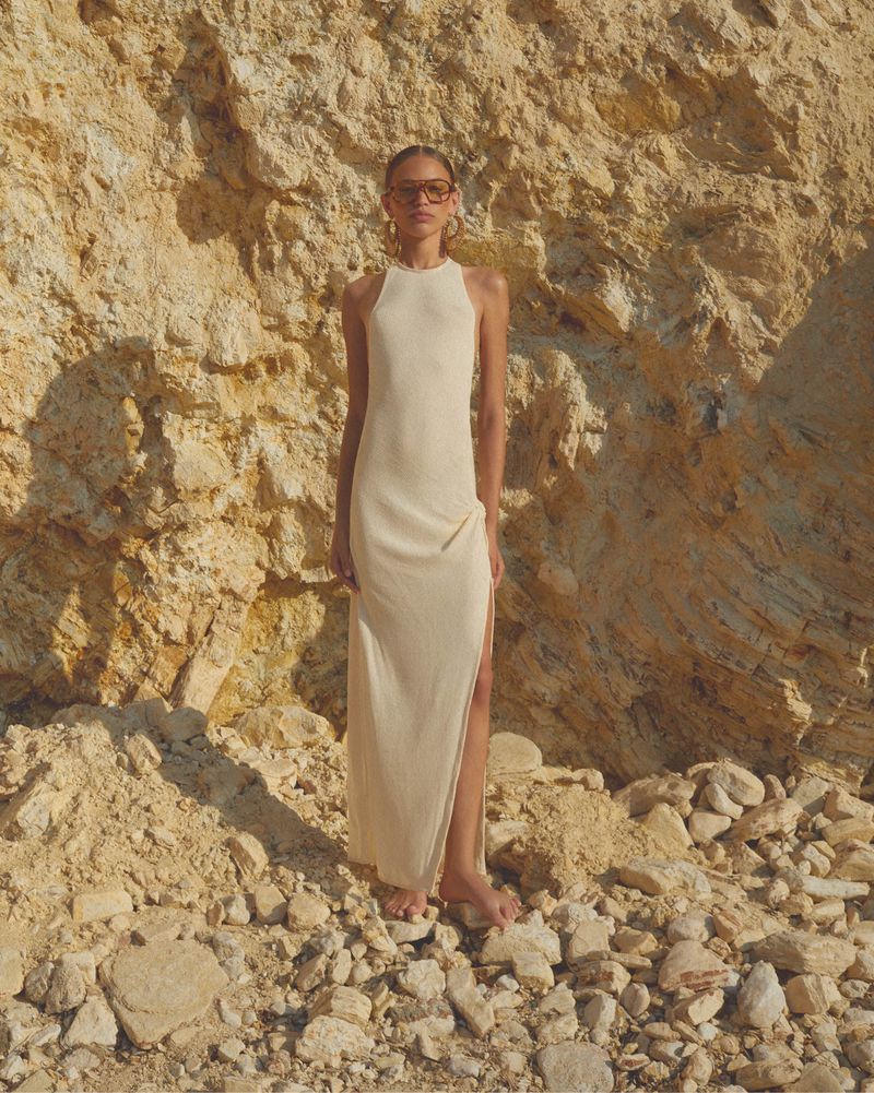 A woman in a cream dress and sunglasses poses against a rock formation, bathed in warm light. The dress features a thigh-high slit.