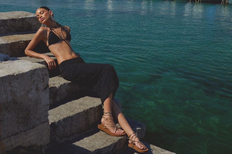 An adult female model poses on stone steps wearing a black bikini and skirt next to teal-blue water in an outdoor environment.