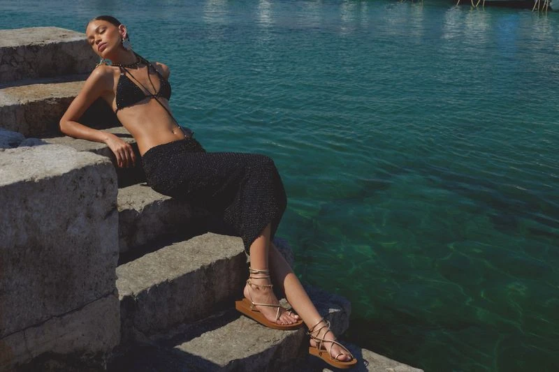 An adult female model poses on stone steps wearing a black bikini and skirt next to teal-blue water in an outdoor environment.