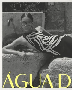 Model Maria Araujo reclines on a rustic stone fountain wearing sunglasses and a patterned black dress in a black and white shot.